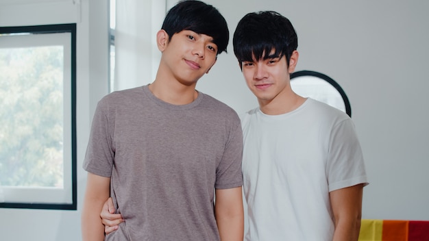 Retrato joven pareja gay asiática se siente feliz sonriendo en casa. Los hombres asiáticos LGBTQ relajan una sonrisa dentuda mirando a la cámara mientras se abrazan en la sala de estar en casa por la mañana.