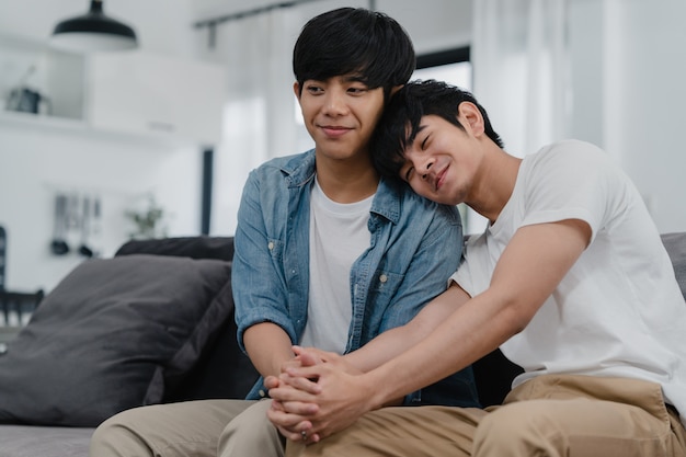 Retrato joven pareja gay asiática se siente feliz sonriendo en casa. Los hombres asiáticos LGBTQ relajan una sonrisa dentuda mirando a la cámara mientras está acostado en el sofá en la sala de estar en casa por la mañana.