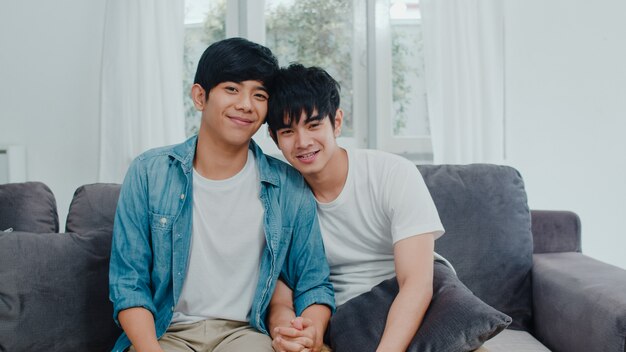 Retrato joven pareja gay asiática se siente feliz sonriendo en casa. Los hombres asiáticos LGBTQ relajan una sonrisa dentuda mirando a la cámara mientras está acostado en el sofá en la sala de estar en casa por la mañana.