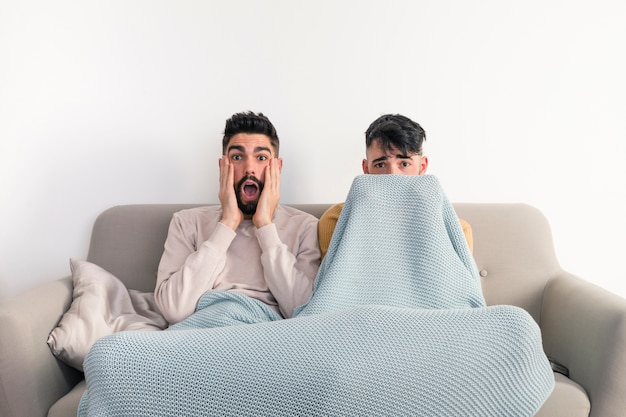 Retrato de joven pareja gay sentada en el sofá viendo una película de terror en la televisión contra una pared blanca
