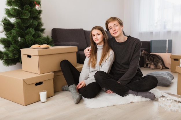 Retrato de una joven pareja hermosa sentada en el suelo en casa y felizmente mirando a la cámara con un gato en el fondo