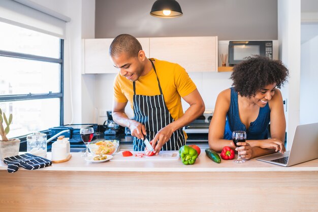 Retrato de joven pareja latina usando un portátil mientras cocina en la cocina de casa. Concepto de relación, cocinero y estilo de vida.