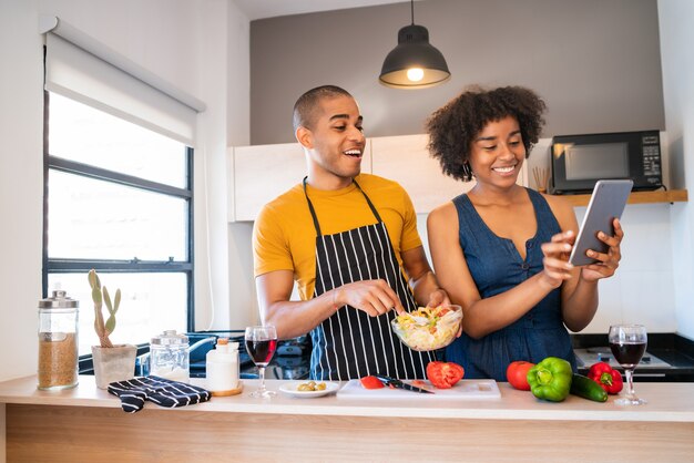 Retrato de joven pareja latina usando una tableta digital y sonriendo mientras cocina en la cocina de casa. Concepto de relación, cocinero y estilo de vida.