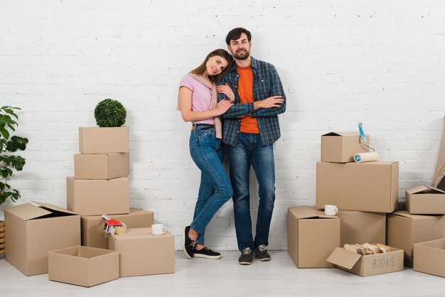 Retrato de joven pareja de pie contra la pared blanca con nuevas cajas de cartón en casa nueva