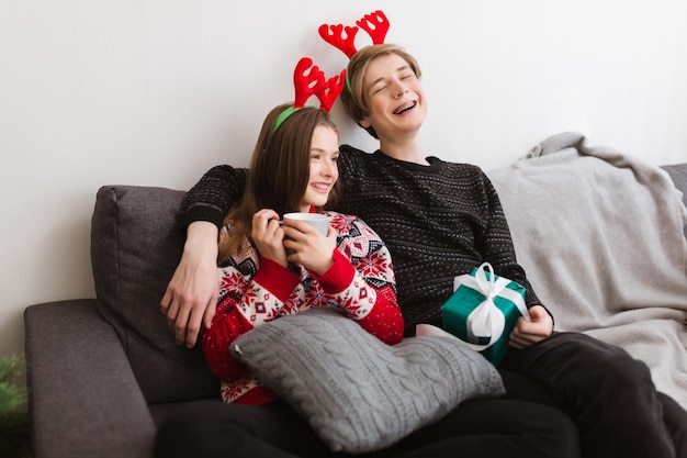 Retrato de una joven pareja sonriente sentada en un sofá en casa y usando cuernos de ciervo mientras pasan tiempo juntos con alegría