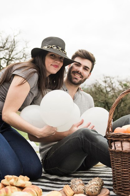 Retrato de la joven pareja sonriente sosteniendo globos blancos en picnic