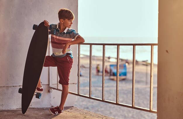 Retrato de un joven patinador vestido con camiseta y pantalones cortos apoyado en una barandilla y sostiene una patineta contra el fondo de una costa al atardecer.