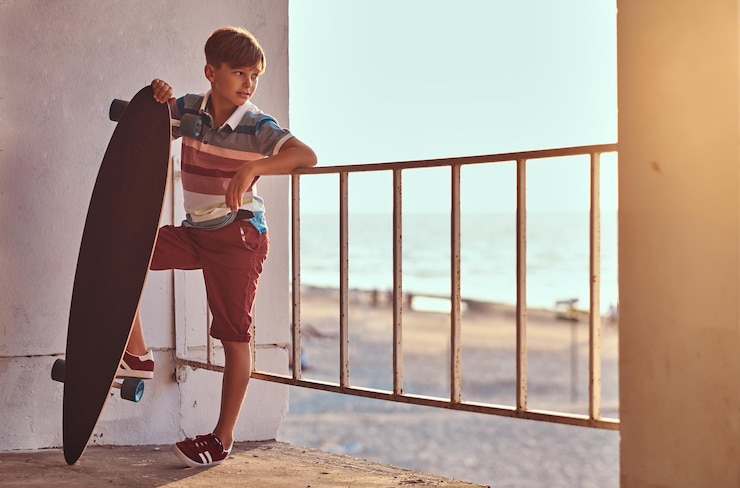 Foto gratuito retrato de un joven patinador vestido con camiseta y pantalones cortos apoyado en una barandilla y sostiene una patineta contra el fondo de una costa al atardecer.