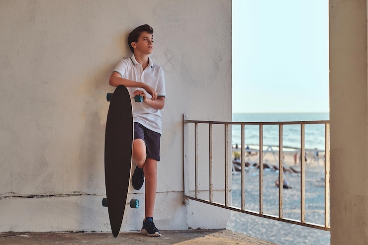 Foto gratuito retrato de un joven patinador vestido con camiseta y pantalones cortos apoyado en una pared y sostiene una patineta contra el fondo de una costa.