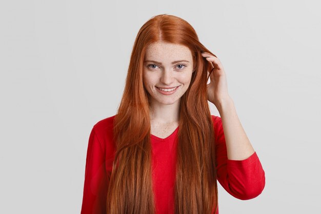 Retrato de joven pelirroja con cabello largo, cara pecosa, sonrisa agradable, toca el cabello