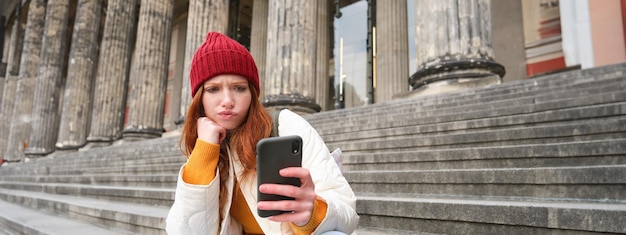 Foto gratuita retrato de una joven pelirroja con cara complicada sentada en las escaleras de la calle con sombrero rojo