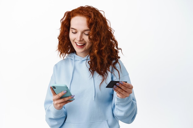 Retrato de una joven pelirroja que paga la compra con un teléfono inteligente y una tarjeta de crédito de plástico mirando los dígitos de la tarjeta de inserción del teléfono móvil con fondo blanco