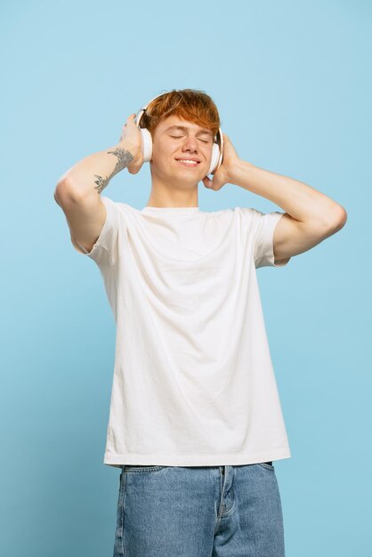 Retrato de un joven pelirrojo escuchando alegremente música en auriculares aislados sobre el fondo azul del estudio