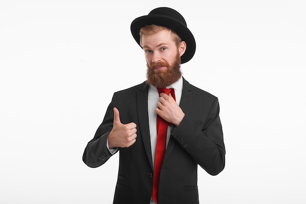 Retrato de un joven pelirrojo con estilo con barba larga recortada posando con ropa elegante de moda, mostrando los pulgares hacia arriba como señal de aprobación, yendo a comprar este traje y sombrero