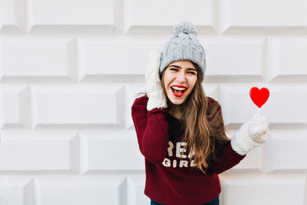 Retrato joven con pelo largo en gorro de punto y suéter marsala con piruleta de corazón rojo en pared gris. Ella usa guantes blancos cálidos, riendo.