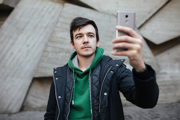 Retrato de un joven de pelo oscuro con chaqueta negra parado en la calle con teléfono móvil en la mano y haciendo fotos en la cámara frontal. Chico con sudadera verde mirando pensativamente en su celular