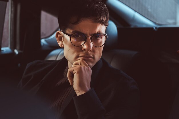 Retrato de un joven pensativo con gafas sentado en el asiento trasero del coche.0