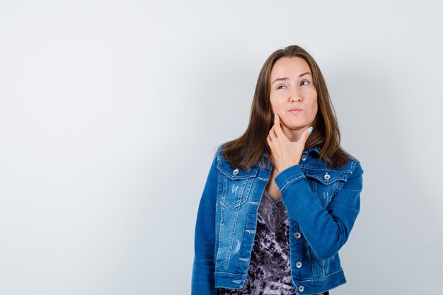 Retrato de joven de pie en pose de pensamiento en blusa, chaqueta de mezclilla y mirando pensativo vista frontal