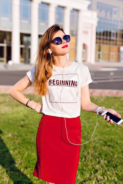 Retrato de una joven de pie sosteniendo un teléfono inteligente y escuchando música en auriculares