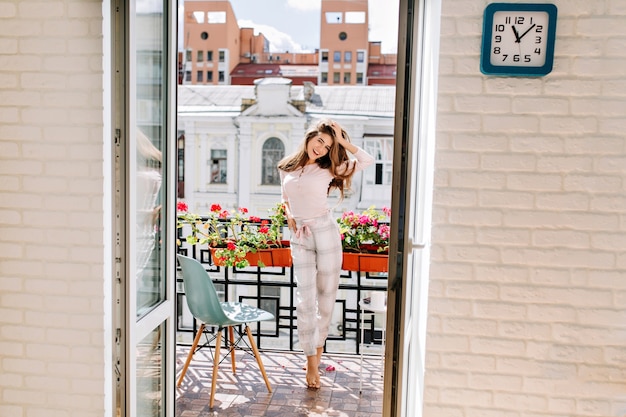 Retrato joven en pijama moviéndose en el balcón de la ciudad en la mañana soleada. Su largo cabello volando con el viento, ella sonriendo.