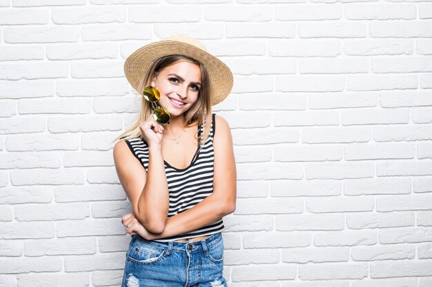 Retrato joven posando con sombrero de paja disfrutando del verano mientras está de pie sobre la pared de ladrillo blanco.