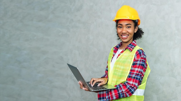 Retrato de una joven profesional de ingeniería civil negra que trabaja en arquitectura usando casco de seguridad para trabajar en un sitio de construcción o en un almacén usando una computadora portátil para trabajar