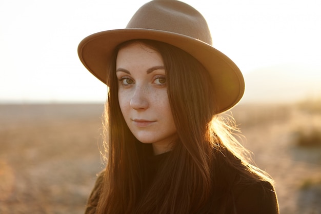 Retrato de joven romántica tierna al aire libre con ojos magnéticos, con sombrero, mirando con tranquilidad y una pequeña sonrisa, caminando en el campo, sintiéndose en armonía con la naturaleza