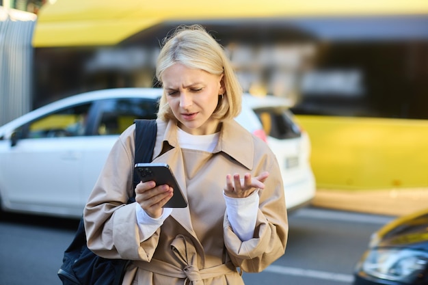 Foto gratuita retrato de una joven rubia confundida de pie en una calle concurrida con coches detrás de ella mirando