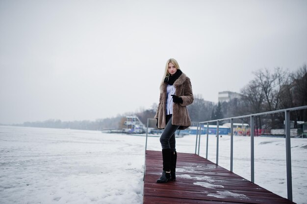 Retrato de una joven rubia elegante con un abrigo de piel en el fondo del muelle río brumoso en el hielo invernal