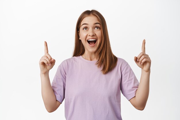 Retrato de una joven rubia emocionada, una chica caucásica señalando y mirando hacia arriba con cara asombrada y feliz, mirando grandes descuentos, pancarta de compras, de pie sobre fondo blanco.