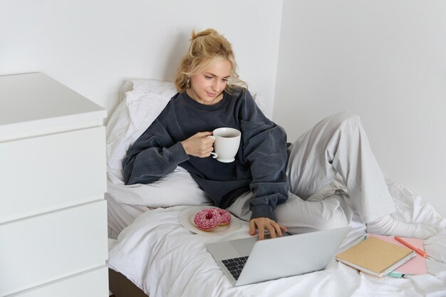 Retrato de una joven rubia hermosa que se siente perezosa en casa acostada en la cama con una taza de rosquilla de té