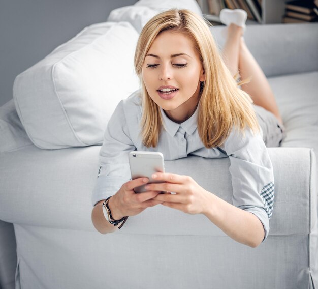 Retrato de una joven rubia tendida en un sofá y enviando SMS en un smartphone.