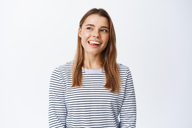 Retrato de una joven sana y feliz sonriendo mostrando la lengua y mirando soñadora en la esquina superior izquierda imaginando algo con cara complacida de fondo blanco