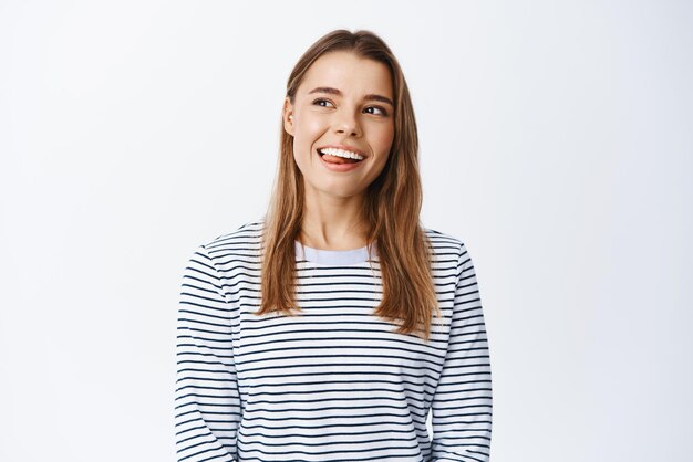 Retrato de una joven sana y feliz sonriendo mostrando la lengua y mirando soñadora en la esquina superior izquierda imaginando algo con cara complacida de fondo blanco