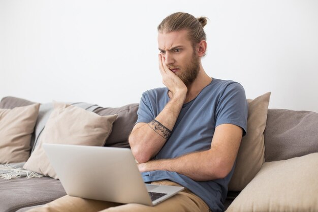 Retrato de un joven sentado en un sofá gris y mirando asombrosamente en una laptop en casa