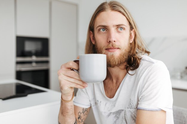 Retrato de un joven sentado con una taza en la mano y mirando soñadoramente a un lado en la cocina de casa