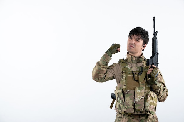 Retrato de joven soldado con ametralladora en camuflaje en pared blanca