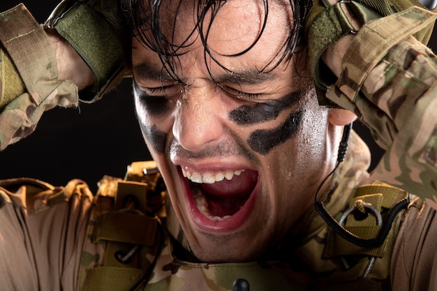 Retrato de joven soldado en camuflaje gritando de dolor en la pared negra