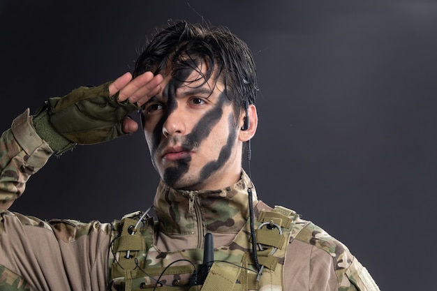 Retrato de joven soldado de camuflaje saludando sobre una pared oscura