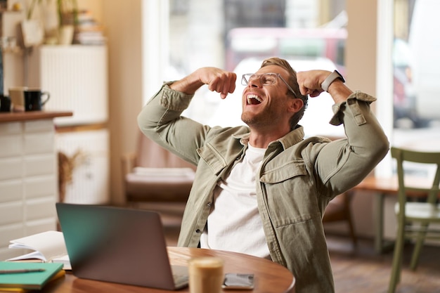 Foto gratuita retrato de un joven sonriente y contento gritando de alegría sentado en un café frente a una computadora portátil