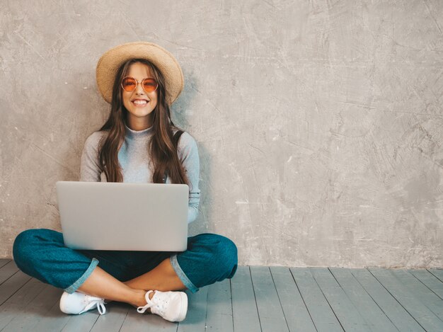 Retrato de joven sonriente creativa en gafas de sol. Hermosa niña sentada en el piso cerca de la pared gris.