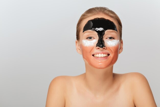 Retrato de una joven sonriente con diferentes máscaras cosméticas en la cara mirando felizmente a la cámara sobre un fondo gris aislado