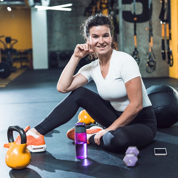 Retrato de una joven sonriente con equipos de ejercicio; celular y botella de agua en el piso