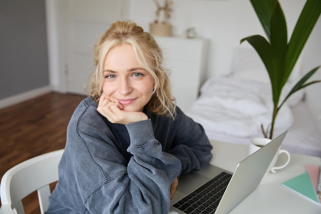 Foto gratuita retrato de una joven sonriente estudiante sentada en su habitación con una computadora portátil mirando con cariño