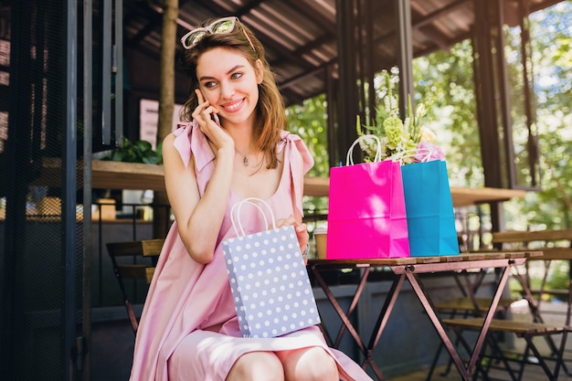 Retrato de joven sonriente feliz atractiva mujer sentada en la cafetería hablando por teléfono con bolsas de la compra, traje de moda de verano, estilo hipster, vestido de algodón rosa, ropa de moda