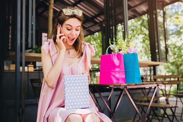 Retrato de joven sonriente feliz atractiva mujer sentada en la cafetería hablando por teléfono con bolsas de compras, traje de moda de verano, estilo hipster, vestido de algodón rosa, cara sorprendida