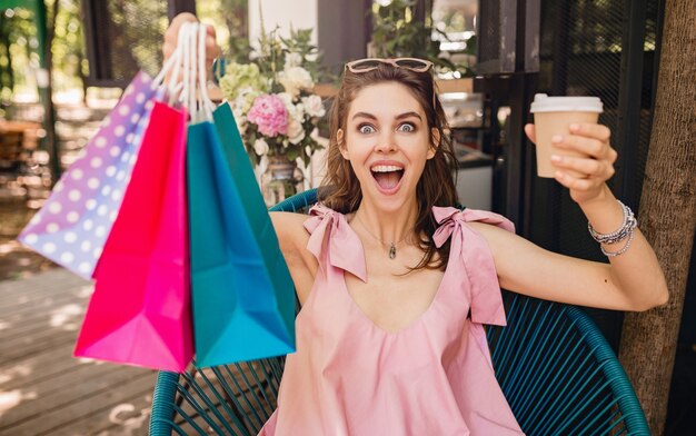 Retrato de joven sonriente feliz mujer bonita con expresión de la cara emocionada sentado en la cafetería con bolsas de compras tomando café, traje de moda de verano, estilo hipster, vestido de algodón rosa, ropa de moda