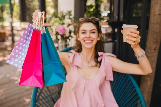 Retrato de joven sonriente feliz mujer bonita con expresión de la cara emocionada sentado en la cafetería con bolsas de compras tomando café, traje de moda de verano, estilo hipster, vestido de algodón rosa, ropa de moda