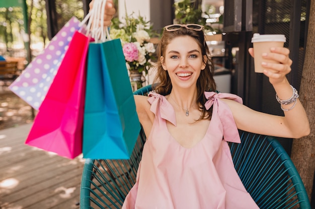 Retrato de joven sonriente mujer bonita feliz con expresión de la cara emocionada sentado en la cafetería con bolsas de compras tomando café, traje de moda de verano, vestido de algodón rosa, ropa de moda