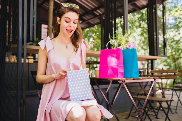 Retrato de joven sonriente mujer bonita feliz con expresión de la cara sorprendida sentado en la cafetería con bolsas de compras, traje de moda de verano, vestido de algodón rosa, ropa de moda